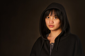Fototapeta premium Mysterious portrait of a young Asian woman in a black hoodie and sweatshirt with a dark background