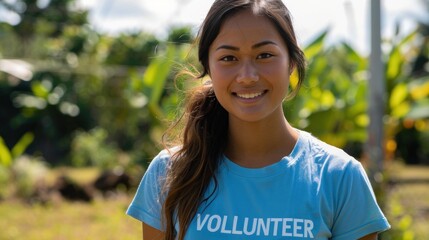 Smiling young woman in blue volunteer shirt standing in front of green plants and trees with a bright and sunny day in the background.