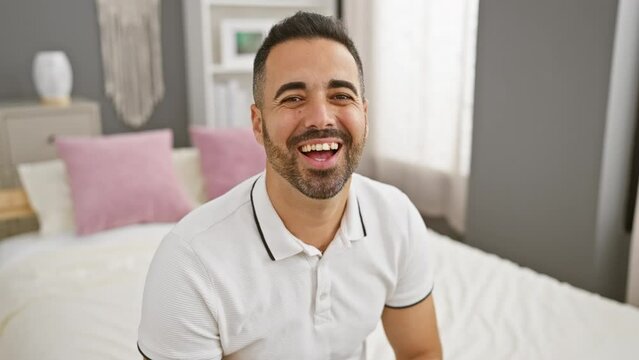 Cheerful young hispanic man caught winking at camera in seductive blink, sitting on bedroom bed with sexy smile