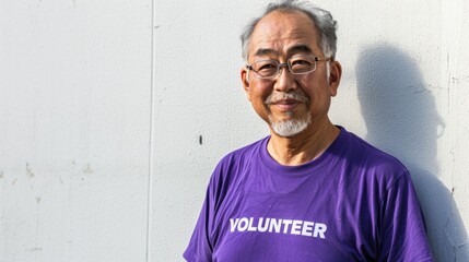 An elderly Asian man with glasses and a beard wearing a purple t-shirt with the word "VOLUNTEER" on it standing against a white wall with a smile on his face.
