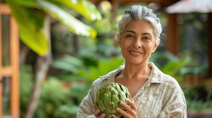 A woman with gray hair smiling holding a large green artichoke surrounded by lush greenery.