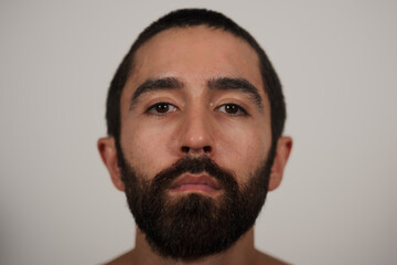 A studio portrait of a multiracial Jewish, Asian, and White man looking neutrally at the camera. His blank expression and shirtless pose highlight his strong eyebrows, dark beard, and dark hair.