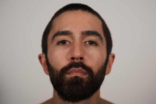 A studio portrait of a multiracial Jewish, Asian, and White man looking neutrally at the camera. His blank expression and shirtless pose highlight his strong eyebrows, dark beard, and dark hair.