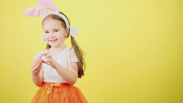 Portrait happy child in bunny ears, looking at camera. Cute little girl pointing on colored egg, posing isolated over yellow color background.