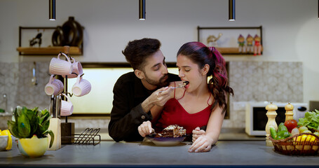 Indian Asian Hindu happy gen z young couple hold spoon eat tasty sweet cake pastry snacks food sitting kitchen enjoy indoor house party adult cute boy girl male female lady talk make fun joy day home