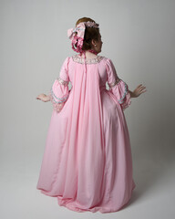 Full length portrait of woman wearing historical French baroque pink gown in style of Marie Antoinette with elegant hairstyle. Standing pose, walking away from the camera, isolated on studio backgroun