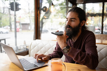 Work from cafe. Young hipster man talking on mobile phone, using voice assistant. Bearded guy with long hair sitting at table with laptop in coffee shop