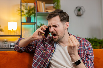Angry young man arguing on smartphone while sitting on couch at home. Quarrel. Caucasian guy talking loudly and shouting into cellphone, maliciously conveying and proving point through gestures.