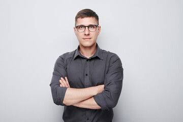 Confident young man in glasses and a gray shirt, arms crossed, standing against a plain background.
