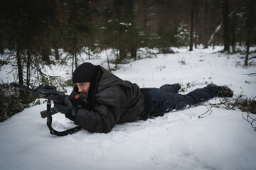 Fototapeta na wymiar A private military man, in a prone position with AK-12 assault rifle in the forest in winter, side view.