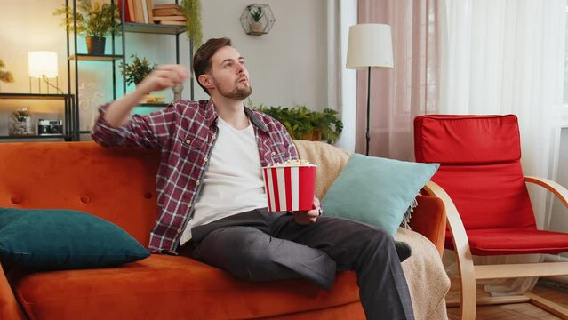 Excited man eating popcorn watching interesting tv serial sport game online social media movie content sitting on sofa in room at home. Happy Caucasian guy in plaid shirt enjoying film during weekend
