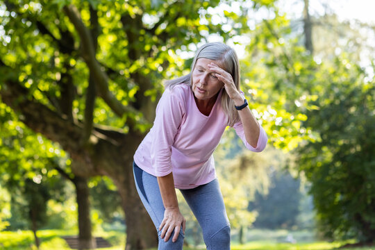 An active senior woman taking a break from running, appears tired, wiping sweat from her forehead in a peaceful park.
