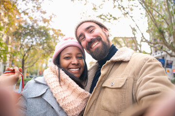 Fototapeta premium Multiethnic smiling young couple with shopping bags making selfie on cell phone outdoors.