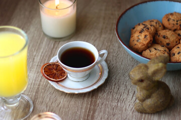 Easter eggs in the basket, bowl of cookies, chocolate pralines, Easter bunny figurine, cups of tea, glasses of juice, flowers and lit candles on the table. Selective focus.