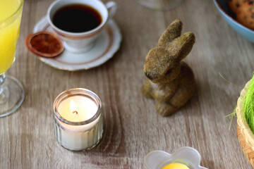 Easter eggs in the basket, bowl of cookies, chocolate pralines, Easter bunny figurine, cups of tea, glasses of juice, flowers and lit candles on the table. Selective focus.