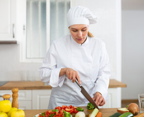 Smiling female chef in white uniform preparing vegetable salad in private kitchen