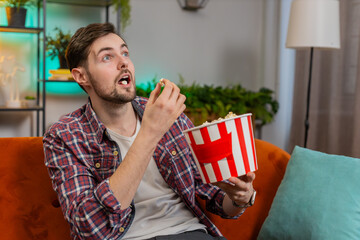 Excited man eating popcorn watching interesting tv serial sport game online social media movie content sitting on sofa in room at home. Happy Caucasian guy in plaid shirt enjoying film during weekends
