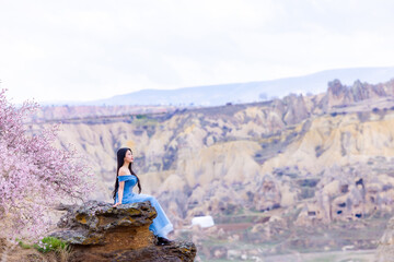 Beautiful woman in blue dress on Turkey rocks mountain.Cheerful woman relax outdoor with beautiful sky and pink flowers in holiday vacation.Woman tourist enjoy amazing mountain view.Travel and freedom