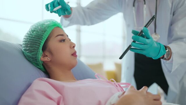 Surgeon explaining and marking young Asian woman face before plastic surgery and showing consulting on tablet before undergoing surgery in surgery clinic. Cosmetic medicine and surgery concept