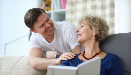 Portrait of happy middle-aged man and his elderly mother having nice conversation in living room. Cheerful senior female reading book on couch. Family relationship concept