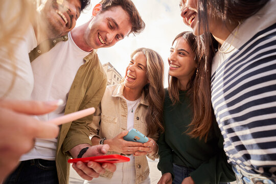A group of young people are standing in a circle, smiling and sharing a cell phone. Happy friends using mobile laughing and having fun togethe