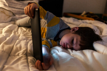 Sleeping young boy lying on his bed watching something on his digital tablet. Light reflection on...