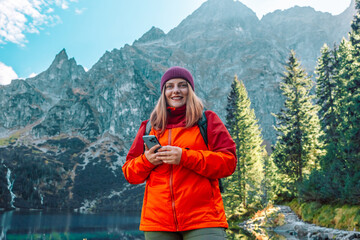 Shot of happy woman looking at the phone with backpack relaxing outdoor with rocky on Morskie Oko lake, hiking mountains. Travel and hobby concept.  - obrazy, fototapety, plakaty