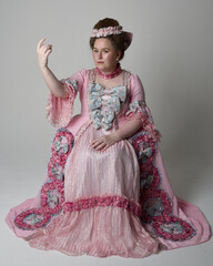 Fun length portrait of female model wearing an opulent pink gown, costume of a historical French baroque nobility, style of Marie Antoinette. Sitting pose on throne. Studio background.