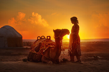 Silhouette of woman in traditional national clothes looks at beautiful sunset in desert next to yurt and camel. - 748029190