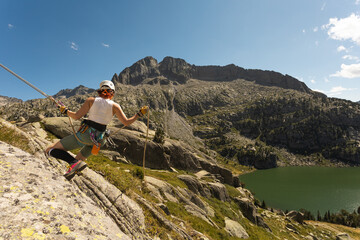 Fototapeta na wymiar Woman climbing on mountain near lake in nature looking at Camara smiling, with blue sky green meadow, wearing helmet yellow jacket with ropes