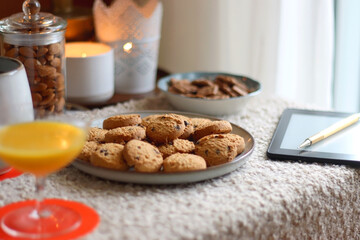 Cup of tea or coffee, stack of books, e-reader, glasses, various cookies, almonds, orange juice and lit candles on the table. Hygge at home. Selective focus.