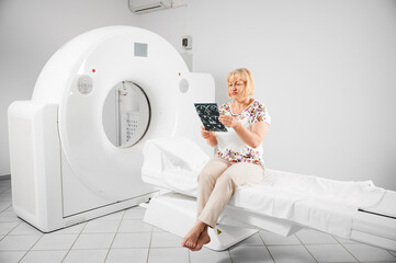 Medical computed tomography or MRI scanner. Patient making MRI, sitting on couchette. Blonde woman holding MRI result, scan, x-ray, looking, examining. Concept of medicine and healthcare.