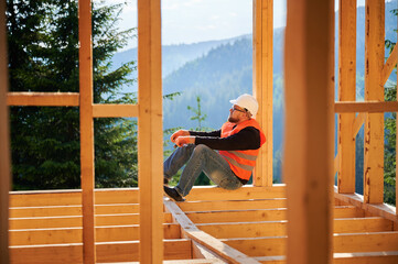 Carpenter constructing wooden frame house near the forest. Bearded man in glasses holding hammer, dressed in protective helmet and orange safety vest. Concept of ecological modern construction.
