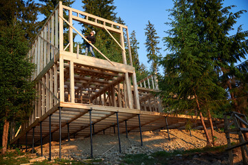 Fototapeta na wymiar Carpenter constructing wooden framed two-story house near the forest. Bearded man with glasses holds hammer, dressed in protective helmet. Concept of environmentally friendly modern construction.