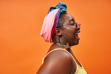 side view of plus size african american woman in headscarf and trendy sunglasses on orange backdrop