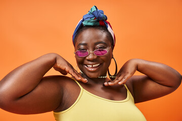 body positive, happy plus size african american woman in headscarf and trendy sunglasses on orange