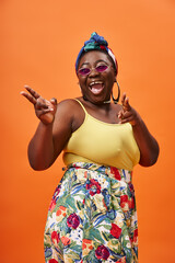 excited african american plus size woman with headscarf and sunglasses posing on orange backdrop
