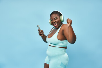 plus size african american woman listening music in headphones from smartphone, blue backdrop