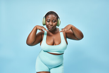 young plus size african american woman in active wear listening music in headphones, blue backdrop