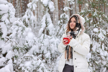 Młoda ładna dziewczyna trzyma w dłoniach czerwony kubek z gorącą herbatą. Spaceruje w mroźny zimowy dzień po lesie. - obrazy, fototapety, plakaty