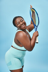 excited plus size dark skinned woman holding tennis racket on blue backdrop, body positive and sport