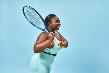 radiant plus size dark skinned woman holding tennis racket on blue backdrop, body positive and sport