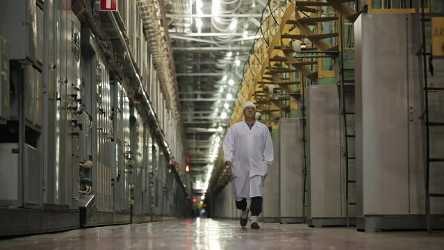 Engineer Is Going Forward To Camera At Factory For Enriched Uranium Manufacturing. Engineer At Work. Long Passage Among Straight Rows Of Bars And Equipment. Walking Engineer At Electrochemical Plant.