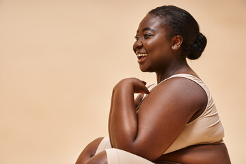 plus size african american model  laughing and sitting against beige backdrop, body positive