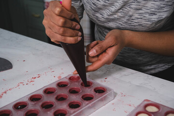 A chef or chocolatier prepares sweet chocolates at home. Close-up of the chocolatier's hands pouring chocolate into the molds. The chef's pastry bag pours hot melted chocolate into a plastic mold.