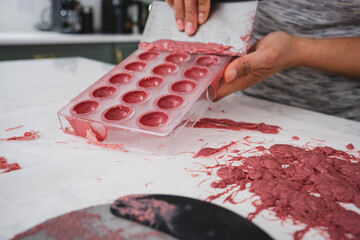 A woman holds in her hands a plastic mold filled with pink chocolate. A woman scrapes excess chocolate from a mold.
Kitchen work surface. Pink chocolate is poured into a plastic mold.