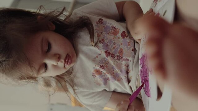 Focused child girl painting a drawing with pink felt-tip pen in a children's coloring book. A cheerful child is engaged in fine art.