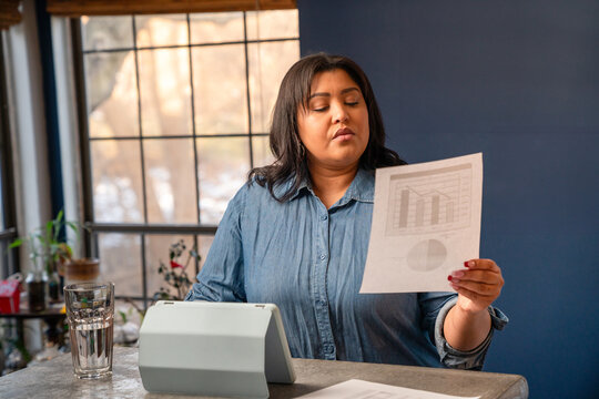 Hispanic Businesswoman Working From Home On A Tablet Looking At A Paper Copy Of A Progress Chart.