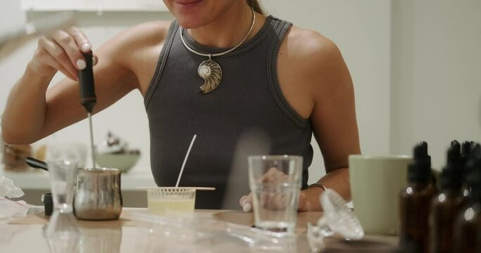 A young woman during a class on making scented candles in a workshop. A woman stirs liquid wax in a jar using a thermometer and smells the resulting aroma. The process of making a handmade candle.
