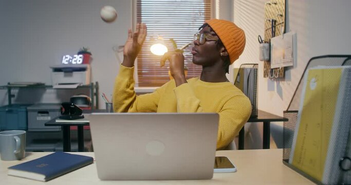 A man with glasses and a knitted hat is playing with a baseball and typing on a laptop sitting at a desk in the office